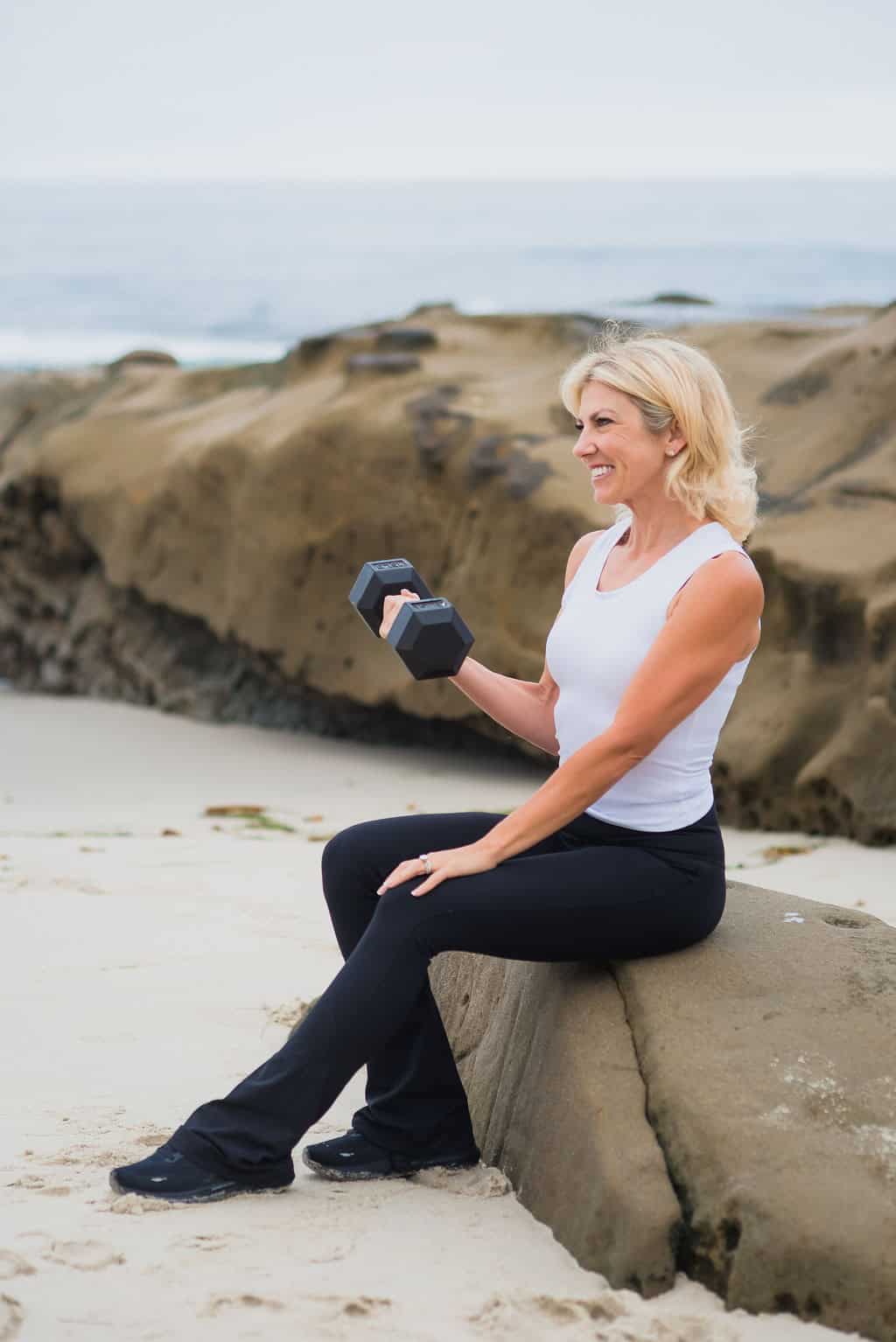 Rose Martin curling a dumbbell while seated on a rock by the shore